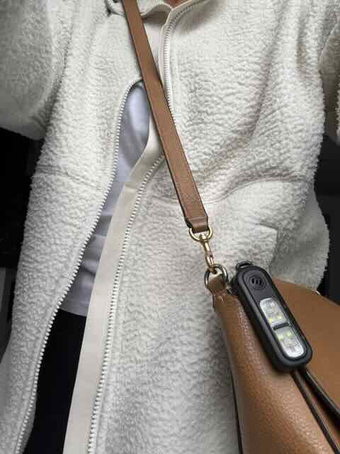 Close-up of a person wearing a light fleece jacket with a brown leather handbag and the personal safety alarm shown clipped onto the bag.